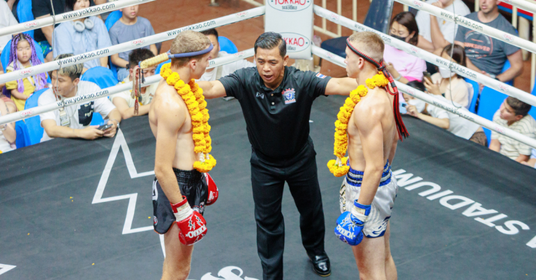 A referee stands before two men in a boxing ring, both prepared for a Muay Thai match, showcasing their athleticism