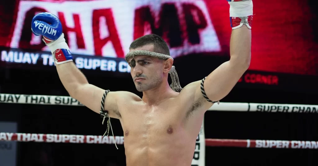 A shirtless Muay Thai fighter in boxing gloves raises his arms in a victorious pose, showcasing strength and determination.